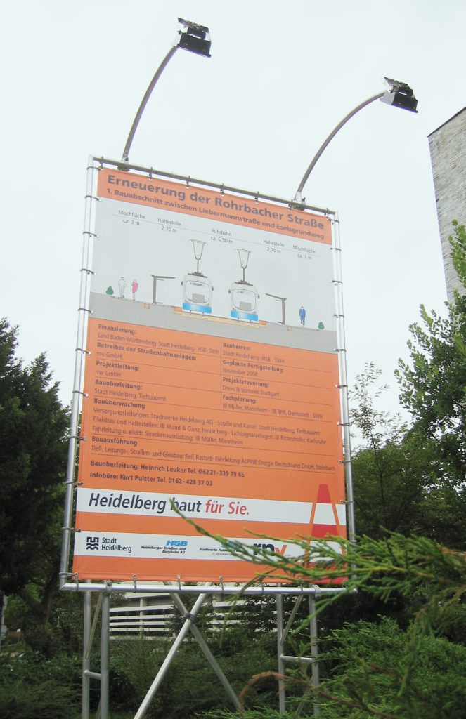 Ein großes Baustellenschild mit großen Strahlern für die nächtliche Beleuchtung. Heidelberg baut für Sie. Erneuerung der Rohrbacher Straße.